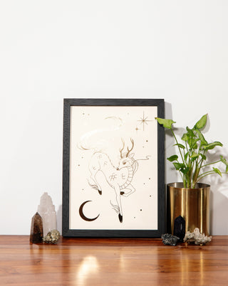 A framed gold foil art print on a wooden table with crystals and pot plant.