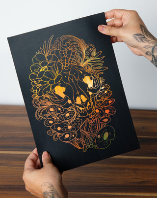 A tattooed hand holding an art print in gold foil of a skull and mushrooms.