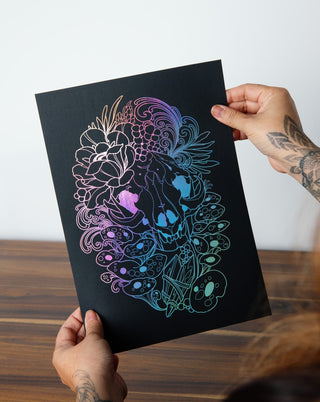 A person holding up an art print in holographic silver foil of a skull and mushrooms.