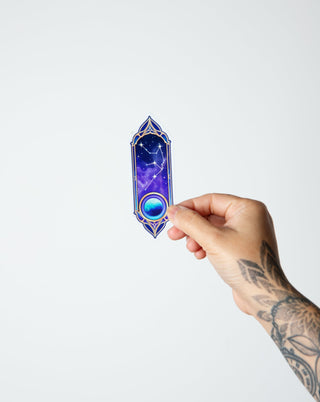 A tattooed hand holding a aquarius zodiac constellation holographic sticker with ruling planet of Uranus in purple and blue.