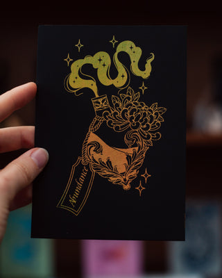 Hand holding a black abundance a5 mini print with gold design against a blurred background