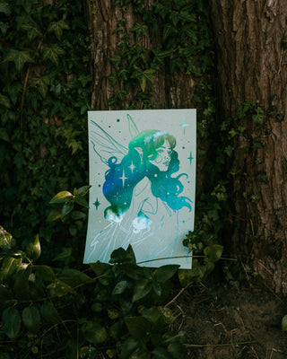 Illustration of a fairy with green hair and wings on a white card, placed among leaves and tree bark.
