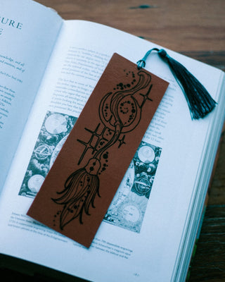 A brown bookmark with black foil witches broomstick pattern and a black tassel, resting on an open book on a dark wooden table.