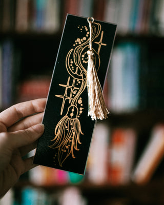 A hand holds a black bookmark featuring an intricate gold foil design of a broomstick with a matching gold tassel, set against a blurred background of bookshelves.