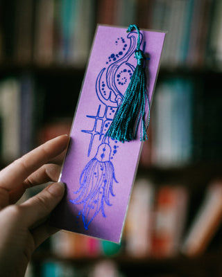 A hand holds a lavender bookmark featuring a witches broomstick with a teal tassel hanging from the top, set against a blurred background of bookshelves.