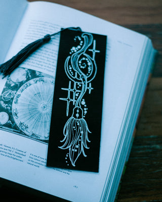 A black bookmark with silver foil broomstick illustration and a black tassel, resting on an open book on a wooden table