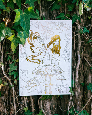 A white art print featuring a gold foil line drawing of a fairy sitting on a mushroom, displayed against a background of green ivy and tree bark.