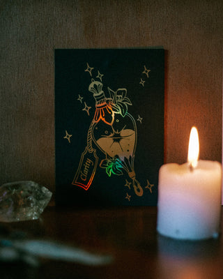 A gold potion A6 card print depicting a 'clarity' bottle illustration with gold foil stars, styled on a wooden shelf next to a lit white candle.