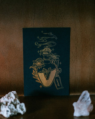 A black art print featuring a gold illustration of a potion bottle, flowers, and smoke, styled on a dark wooden surface with white crystals in the foreground.