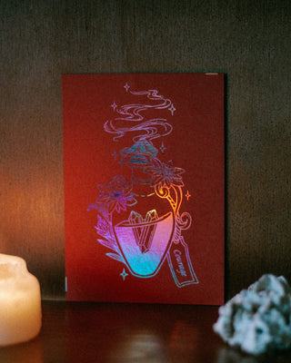 A red mini A6 print with a stylized iridescent potion bottle illustration in pink tones, standing on a dark surface beside a lit candle and a small white crystal.