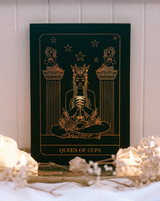 'Queen of Cups' art print in gold illustrations on a dark green background, placed on a surface with candles and crystals.