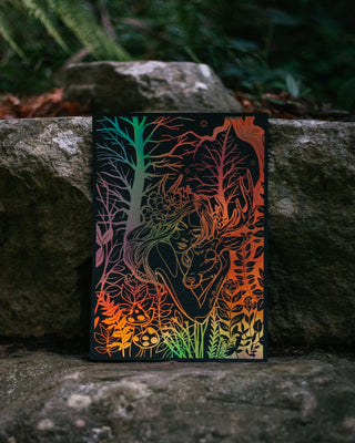 An art print featuring a stylized black silhouette of a woman hugging a deer surrounded by botanical and trees linework, leaning against large outdoor rocks with blurred greenery in the background.