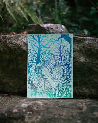 A art print featuring a stylized line-art illustration of a woman among trees and foliage hugging a deer, rendered in shades of blue and teal on a pale mint background. The print is propped against a large stone outdoors with blurred greenery behind it.