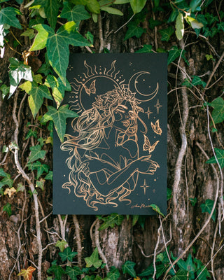 A black art print featuring a gold foil drawing of a fairy with a crescent moon, sun, and butterflies, displayed against a real tree trunk covered in green ivy.