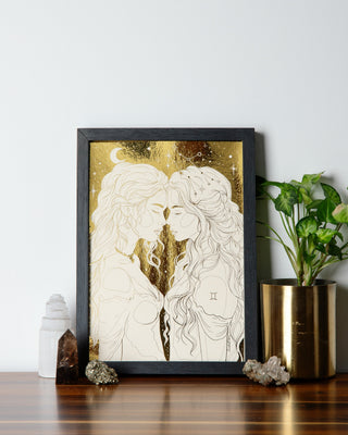A framed art print depicting two feminine figures in a warm color palette with celestial details, displayed on a wooden surface with decorative objects around it.
