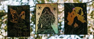 Three artistic prints of women in nature settings hanging on a tree branch with a blurred forest background.