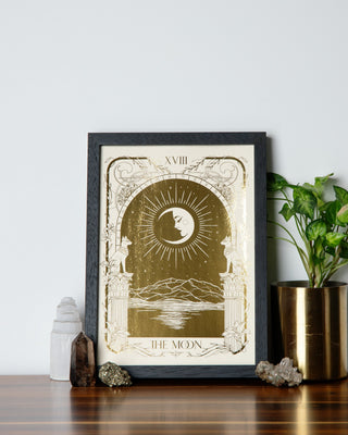 A framed gold Tarot Moon art print surrounded with crystals and pot plant on a wooden table.