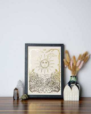 A framed print titled 'The Sun' with solar motifs and a sun-kissed shimmer, displayed on a wooden surface with decorative items.