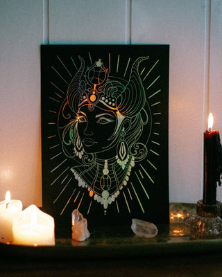 A black art print or canvas featuring an ornate white line drawing of Hathor with radiating lines and decorative jewellery, displayed on a shelf beside lit candles and crystals against a panelled wall.