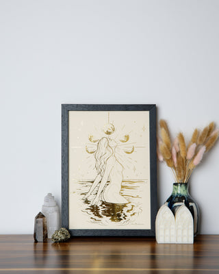 An art print featuring a stylized, ethereal female figure with a fluid, graceful pose, set against a moonlit water background, framed in a black frame and displayed on a wooden surface with decorative items around it.