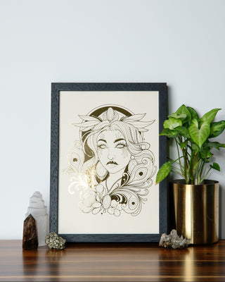 A framed art print featuring a detailed black line art illustration of a woman's face with floral and leaf motifs, styled on a wooden shelf next to a potted green plant and crystals.