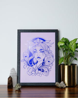 A framed art print featuring a blue line-art illustration of a woman's face with ornate hair details on a light lavender background, styled on a wooden surface next to a potted green plant and crystals.
