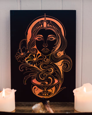 Decorative wall art with goddess Hestia and floral elements on a black background, placed on a table with candles.