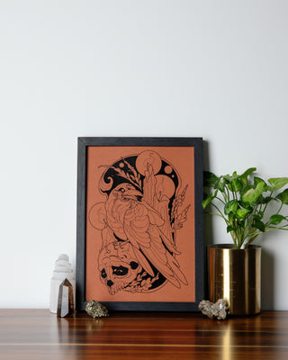 A beautiful framed art print with a raven and skull on it on a table with crystals and pot plant nearby.