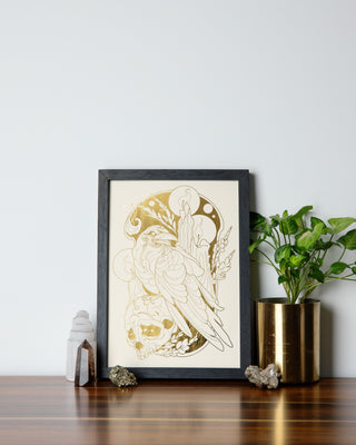 A beautifully framed art print in gold foil and cream on a table with crystals and pot plant.