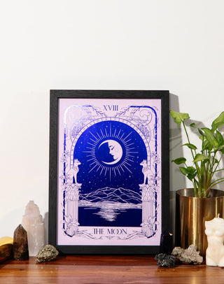 A framed tarot art print titled 'The Moon' (XVIII) featuring a deep blue and lavender design, styled on a wooden shelf next to a potted plant, and crystals against a white wall.