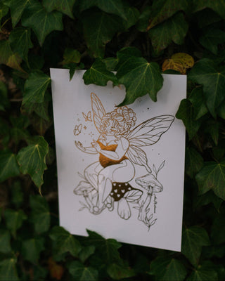 A white art print featuring a delicate line drawing of a fairy with gold accents, displayed against a background of green ivy leaves.