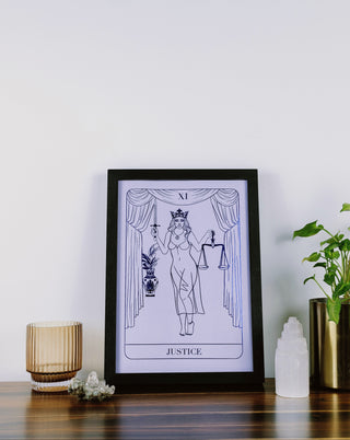 A framed print titled 'Justice' from tarot series, depicting a figure holding a sword and scales, placed on a wooden surface with decorative items around it.