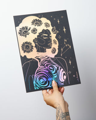 A hand with tattoos holds up an art print featuring a stylized black silhouette of a woman with flowers in her hair and a colorful iridescent spiral pattern on her chest, against a plain light gray wall.