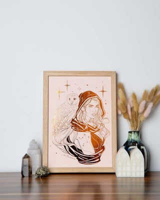 A framed illustration of a Libra zodiac sign, featuring a female figure with an owl, set against a wooden frame on a table with decorative items.