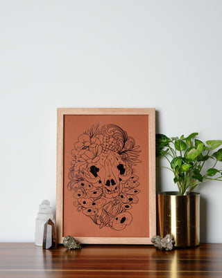 A beautifully framed art print with an animal skull and mushrooms on a table with crystals and pot plant.