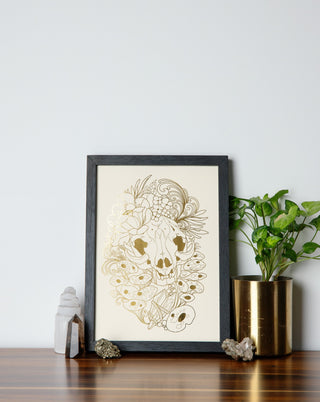 A framed art print in gold foil on a table with crystals and pot plant nearby.