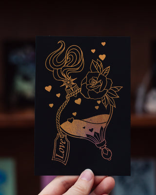 Hand holding a black card with gold artwork of a love potion bottle, rose, and heart on a blurred background.