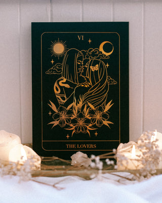 Decorative tarot card titled 'The Lovers' with gold illustrations on a dark background, placed on a white surface with candles.