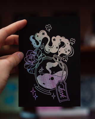 Hand holding a black card with a colorful design of a luck potion bottle and flowers on a blurred background.