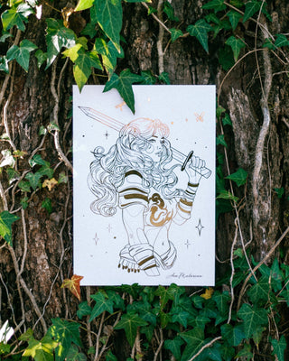 An A4 art print featuring gold line foil illustration of a female warrior with a sword, styled in armour, displayed against a tree trunk covered in green ivy.