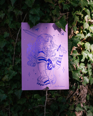 An art print featuring an indigo foil of a female warrior character holding a sword, displayed against a background of green ivy.