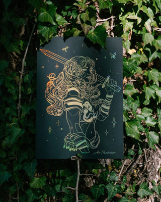 A black art print featuring a fine line-art illustration of a fantasy warrior with a sword, displayed against a background of real green ivy.