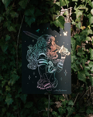 A black art print featuring a female fairy with a sword, printed with holographic silver foil, displayed against a background of green ivy.