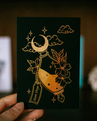 Black card with gold moon design held by a hand against a dark background