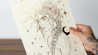 An art print featuring a stylized, graceful feminine figure with a solar system and stars background, held in a person's hand.