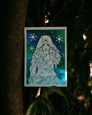 An art print featuring an illustration of a whimsical winter fairy with flowing hair against a teal and blue background with snowflake details, photographed hanging against a dark tree trunk and foliage.