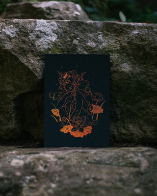 A black art print featuring an intricate copper-orange line drawing of a fantasy creature with lotus flowers, displayed on a rustic stone surface with a blurred natural background.
