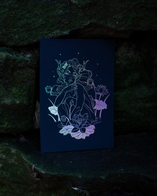 A black art print featuring a stylized line-art illustration of a nymph surrounded by lotus flowers, set against a dark, mossy stone background.
