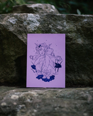 An art print with a lavender background and a indigo line-art illustration of a woman surrounded by lotus flowers, displayed on a textured gray stone surface.
