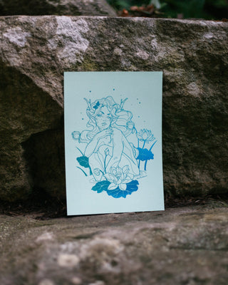 An art print featuring a nymph surrounded by lotus flowers on a pale mint-green background, displayed leaning against a textured gray stone outdoors.